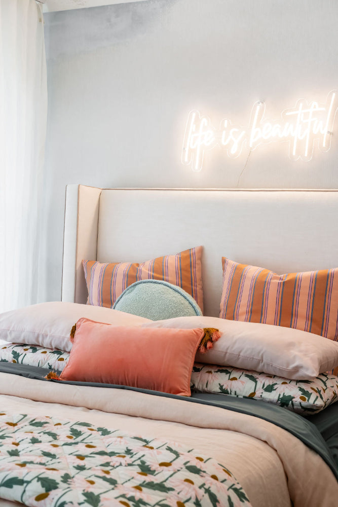 A white neon sign turned on in the daylight, mounted above a bed with beautiful linen