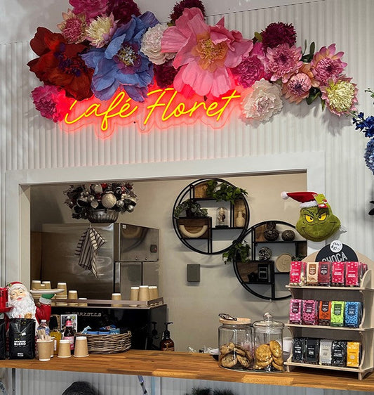 Red script neon sign reading Café Floret above a café counter, under a flower display