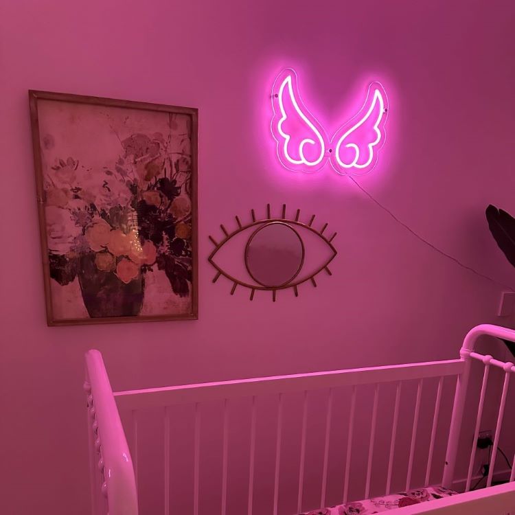 Pink neon angel wings on a nursery wall above a white cot