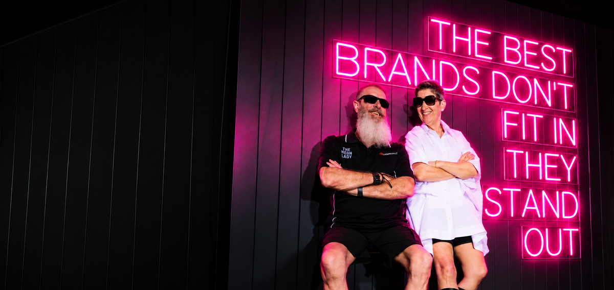 Russell and Shaz from The Neon Lady smiling under a hot pink LED neon sign that reads The best brands don't fit in, they stand out