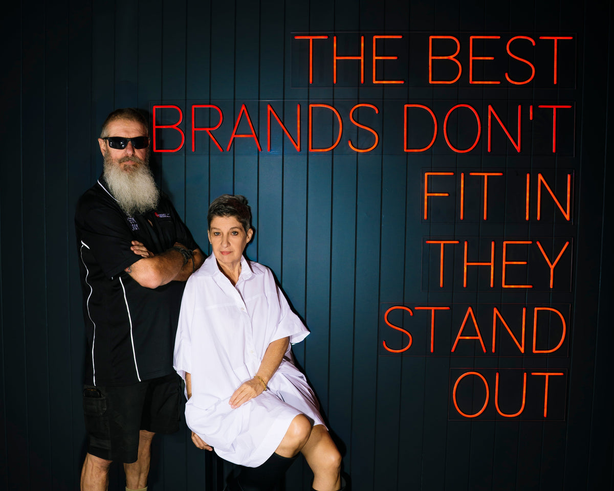 A man with a long white beard and sunglasses stands next to a woman in a white shirt and black boots, posed against a dark paneled wall with large orange text reading 'THE BEST BRANDS DON'T FIT IN THEY STAND OUT'.