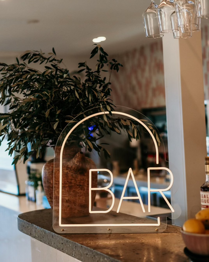 arch shpe warm white neon sign on an concrete bar with small olive tree behind