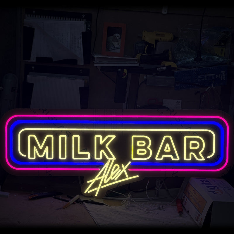 Alex Milk Bar neon sign in yellow with pink and blue borders and an Alex signature, on the workshop bench