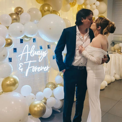 A couple kissing in front of an LED neon sign that reads 'Always and Forever', with a backdrop of white and gold balloons.