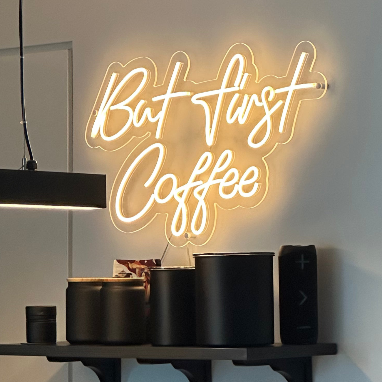 "but first coffee" warm white neon sign above a shelf with black canisters 