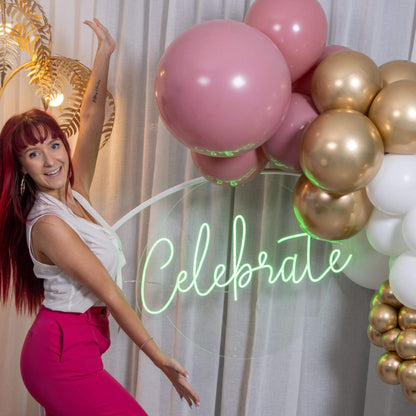 A person standing next to a neon sign that reads 'Celebrate' in a cursive font, surrounded by decorative balloons.