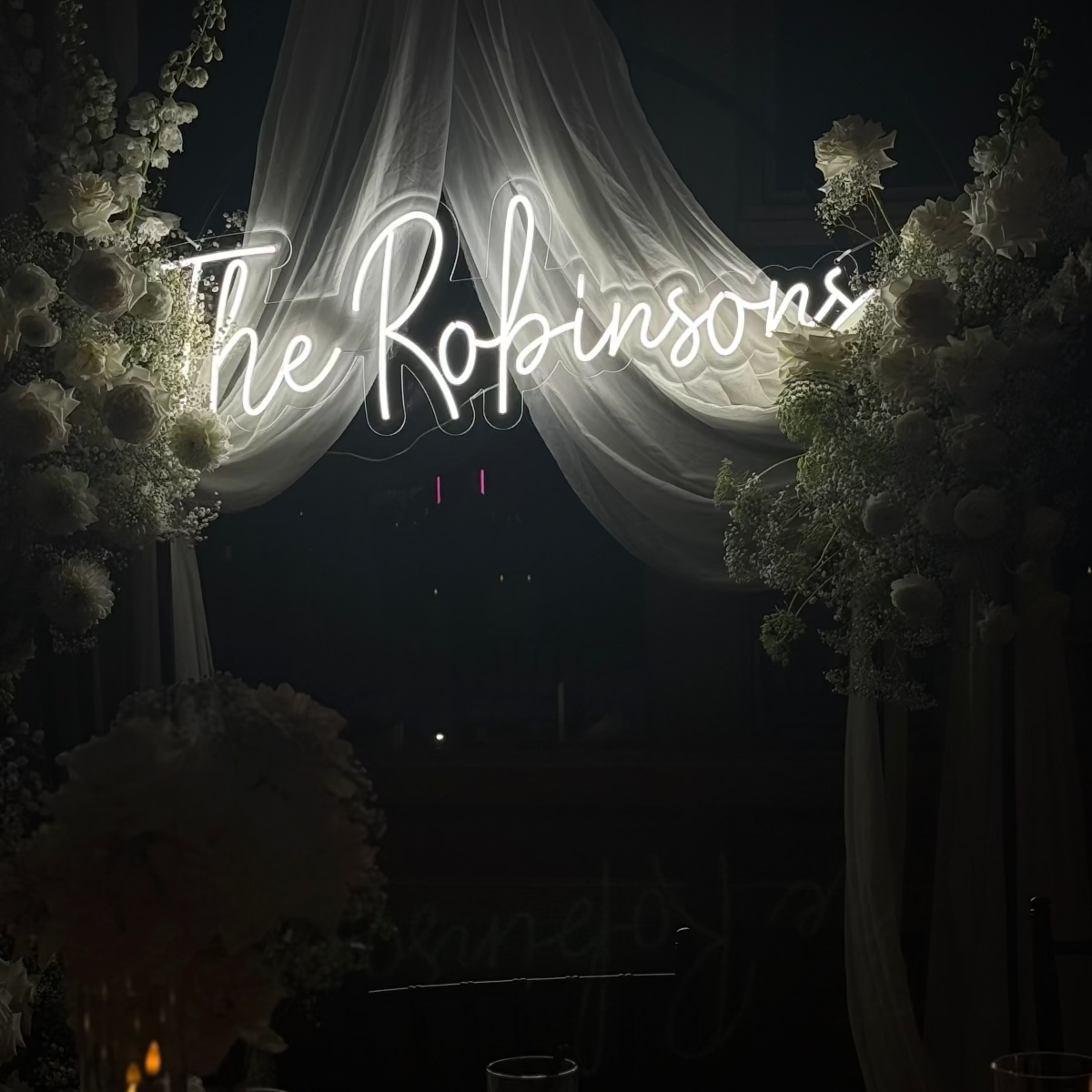 Neon sign reading 'The Robinsons' above a table with candles and flowers at night.