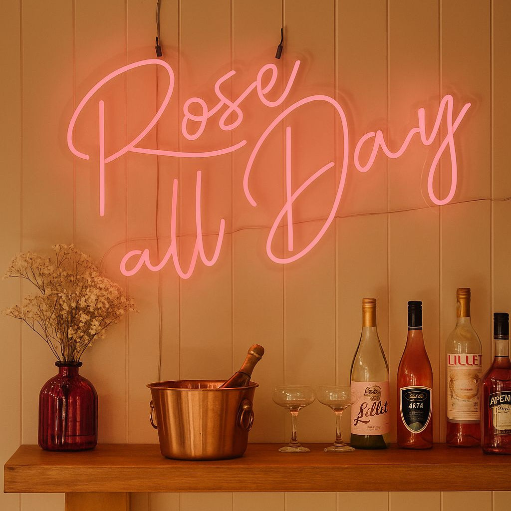 Neon sign reading 'Rose all Day' above a wooden bar with wine bottles and glasses.