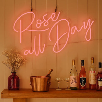 Neon sign reading 'Rose all Day' above a wooden bar with wine bottles and glasses.